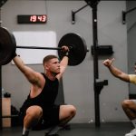 A muscular man performing a weightlifting squat with a coach guiding him indoors.