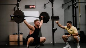 A muscular man performing a weightlifting squat with a coach guiding him indoors.