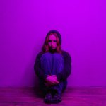 A woman in a hoodie sits alone on the floor, illuminated by purple light indoors.